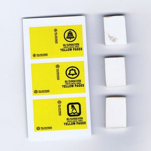 Könnte beinhalten: Drei weiße Aufkleber mit dem Text "Yellow Pages" und dem Logo des Bell Telephone Systems. Das Logo zeigt eine Glocke mit dem Schriftzug "Bell System" darin. Die Aufkleber befinden sich auf einem weißen Blatt Papier.