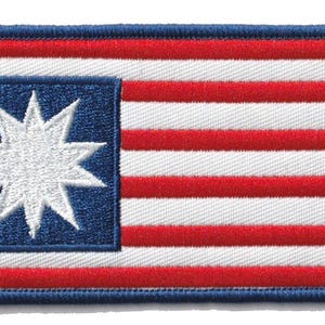 以下が含まれることがあります： 旗のデザインが施された刺繍パッチ。青い縁取りと、白い星型の入った青い正方形があります。パッチの残りの部分は赤と白のストライプです。長方形の形をしています。