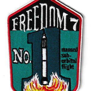 Peut inclure: Écusson brodé en forme de bouclier, avec le texte "FREEDOM 7" en blanc, au-dessus d'un grand chiffre "1". Une fusée décolle des flammes, avec le texte "manned sub-orbital flight". L'écusson a une bordure rouge.