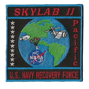 Op de afbeelding: Geborduurde patch met de tekst "Skylab II Pacific" in het rood, met een blauwe rand. De patch bevat een wereldbol met een raket, parachute en NASA-logo. De tekst "U.S. Navy Recovery Force" is ook aanwezig.