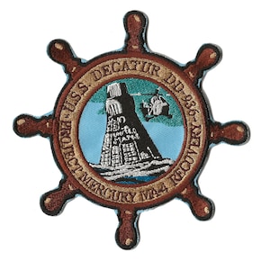 Op de afbeelding: Een bruin geborduurd patch met een scheepswielontwerp. Het midden van het patch heeft een blauwe achtergrond met een witte vuurtoren en een helikopter. De tekst rond de rand luidt "USS DECATUR DDG 36 PROJECT MERCURY 1964 RECOVERY"