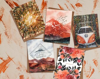 May include: Set of five greeting cards with watercolour illustrations. The cards feature a mountain range, a floral design, a sun rising over a desert landscape, a vintage van, and a forest scene. The cards are all blank inside for writing a message.