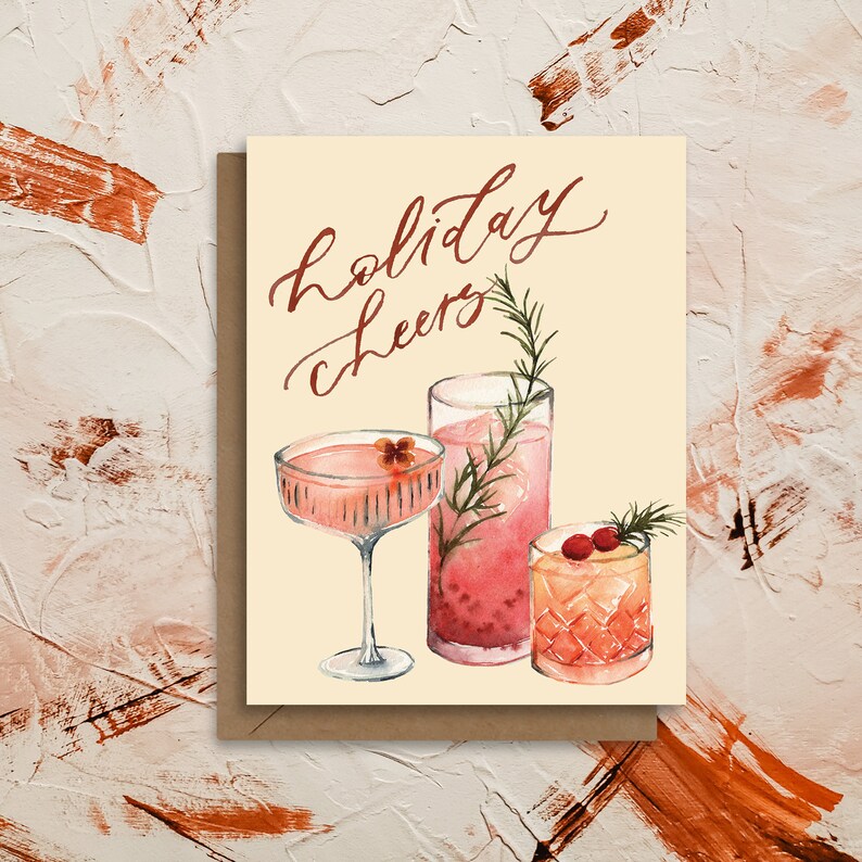 May include: A holiday greeting card with the text "holiday cheers" in red cursive lettering. The card features a watercolor illustration of three pink cocktails with garnishes.