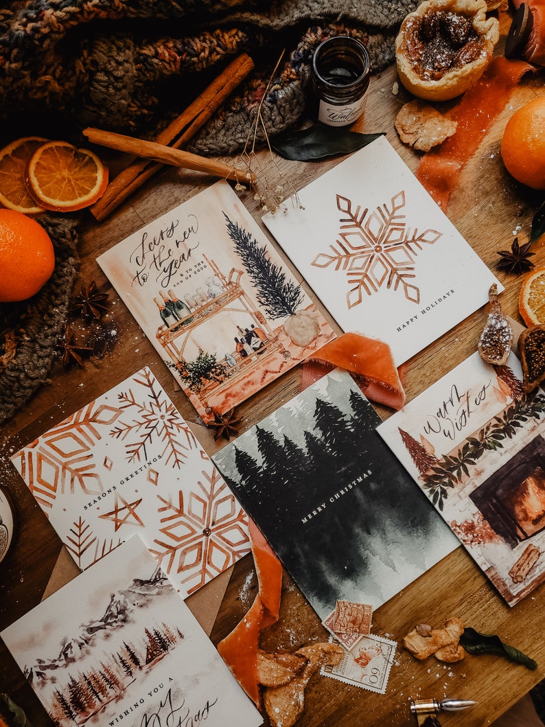 May include: A collection of Christmas cards with various winter themes. The cards feature watercolor illustrations of snowflakes, a forest, a fireplace, and a winter scene with mountains. The cards are all white with gold accents and have handwritten calligraphy.
