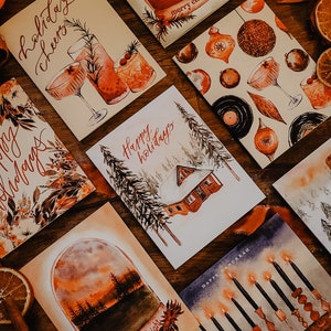 May include: A collection of eight watercolor holiday greeting cards featuring various winter themes. The cards are arranged on a wooden surface with dried orange slices and cinnamon sticks. The cards feature illustrations of a winter cabin, a snowy landscape, a menorah, a Christmas tree, and a winter scene with a glass of wine.