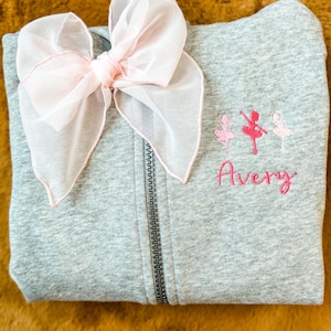 May include: Gray zippered sweatshirt with a pink bow on the front. The sweatshirt has pink embroidered ballerinas and the name "Avery" on the front.