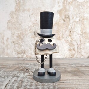 May include: A white ghost figurine with a black top hat, a black mustache, and a monocle. The ghost is wearing black shoes and has a silver chain attached to the monocle.