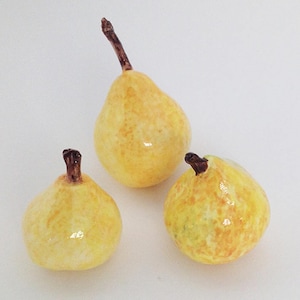 May include: Three yellow ceramic pears with brown stems. The pears are arranged on a white surface.