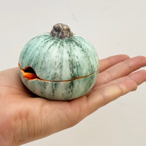 May include: A small, round, ceramic trinket box shaped like a gourd, held in a hand. The box is primarily light green with darker green marbling and a brown stem. The interior is orange. The box is open, revealing the inside.