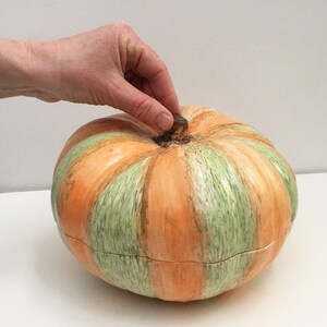 May include: A ceramic pumpkin-shaped serving dish with a green and orange striped pattern. The dish is closed and has a small brown stem on top.