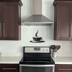 May include: A stainless steel range hood with a black coffee cup and steam design decal above it. The range hood is installed above a black electric stovetop with a white countertop.
