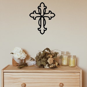 May include: A wooden dresser with three drawers and a black metal cross on the wall above it. The dresser has two round wooden knobs on each drawer. There are white flowers in a brown pot and a bouquet of dried flowers on top of the dresser. There are three glass jars with candles on the dresser.