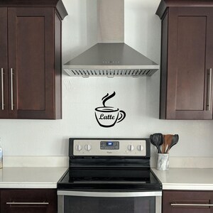 May include: A kitchen with dark brown cabinets, a stainless steel range hood, and a white backsplash with a black coffee cup decal that says "Latte".