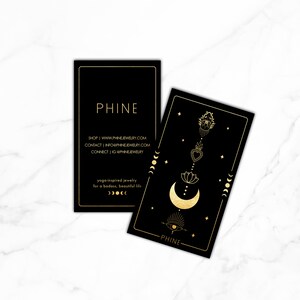 May include: Two black business cards with gold lettering and illustrations. The top card has the text "PHINE" and website information. The bottom card has a gold illustration of a crescent moon, a lotus flower, a heart, and a hand with an eye. The text "PHINE" is at the bottom of the card.