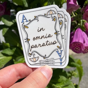 May include: White sticker with the text "in omnia paratus" in a decorative font. The sticker has a playing card design with an umbrella and the letter "A" in the corners. The sticker is held in front of a green and purple floral background.