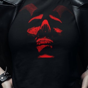 Puede incluir: Camiseta negra con un gráfico de calavera roja. La calavera tiene cuernos y una expresión amenazante.