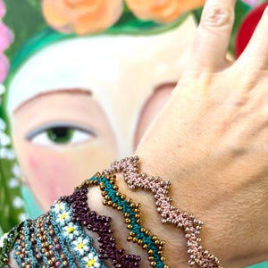 May include: A collection of beaded bracelets in various colors, including teal, purple, and pink. The bracelets feature intricate patterns and gold-colored beads. The background is a painting of a woman with flowers in her hair.