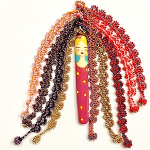 Op de afbeelding: Handgemaakte hanger met een beschilderde houten pop, roze lichaam en geel haar. De pop is omgeven door kralenstrengen in verschillende kleuren, waaronder rood, oranje, goud en paars. De kralen zijn gerangschikt in een bloemmotief.