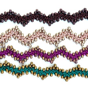 May include: Five beaded zig-zag trim samples in various colors. The colors include dark brown, light pink, purple, and teal. Each trim is adorned with small, gold-colored beads. The trim is likely used for embellishing clothing or craft projects.