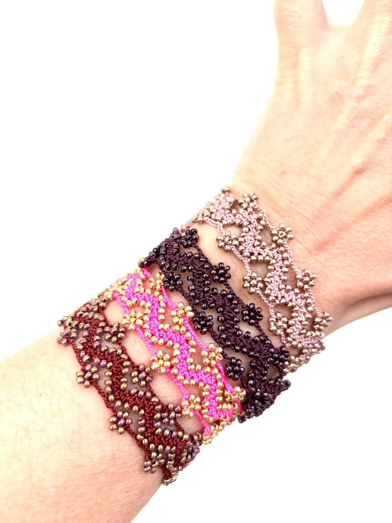 May include: A collection of handmade bracelets in various colors, including burgundy, pink, and mauve. Each bracelet features a delicate floral pattern with gold-colored beads, creating a unique and elegant accessory. The bracelets are displayed on a wrist.