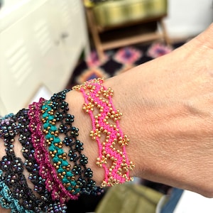 May include: A collection of beaded bracelets in various colors, including black, teal, pink, and purple. The bracelets feature intricate floral patterns and are adorned with gold-colored beads. The bracelets are displayed on a wrist.