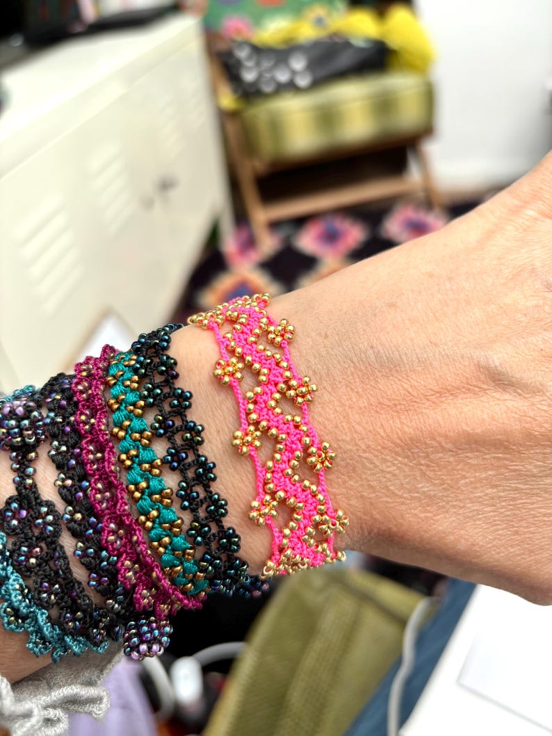 May include: A collection of beaded bracelets in various colors, including black, teal, pink, and purple. The bracelets feature intricate floral patterns and are adorned with gold-colored beads. The bracelets are displayed on a wrist.