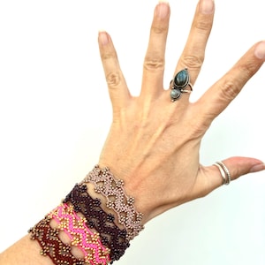 May include: A hand displays several beaded bracelets in shades of pink, brown, and gold. The bracelets have a floral design. A silver ring with a teardrop-shaped stone and a smaller round stone is on a finger.