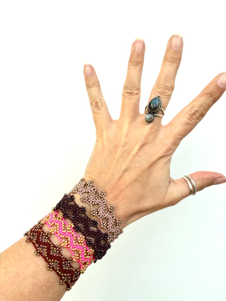 May include: A hand displays several beaded bracelets in shades of pink, brown, and gold. The bracelets have a floral design. A silver ring with a teardrop-shaped stone and a smaller round stone is on a finger.
