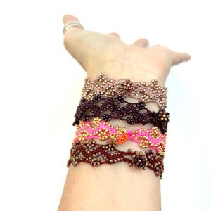 May include: A collection of beaded bracelets in various colors, including pink, brown, and gold. The bracelets are intricately designed with floral patterns and are stacked on a wrist. The background is plain white.