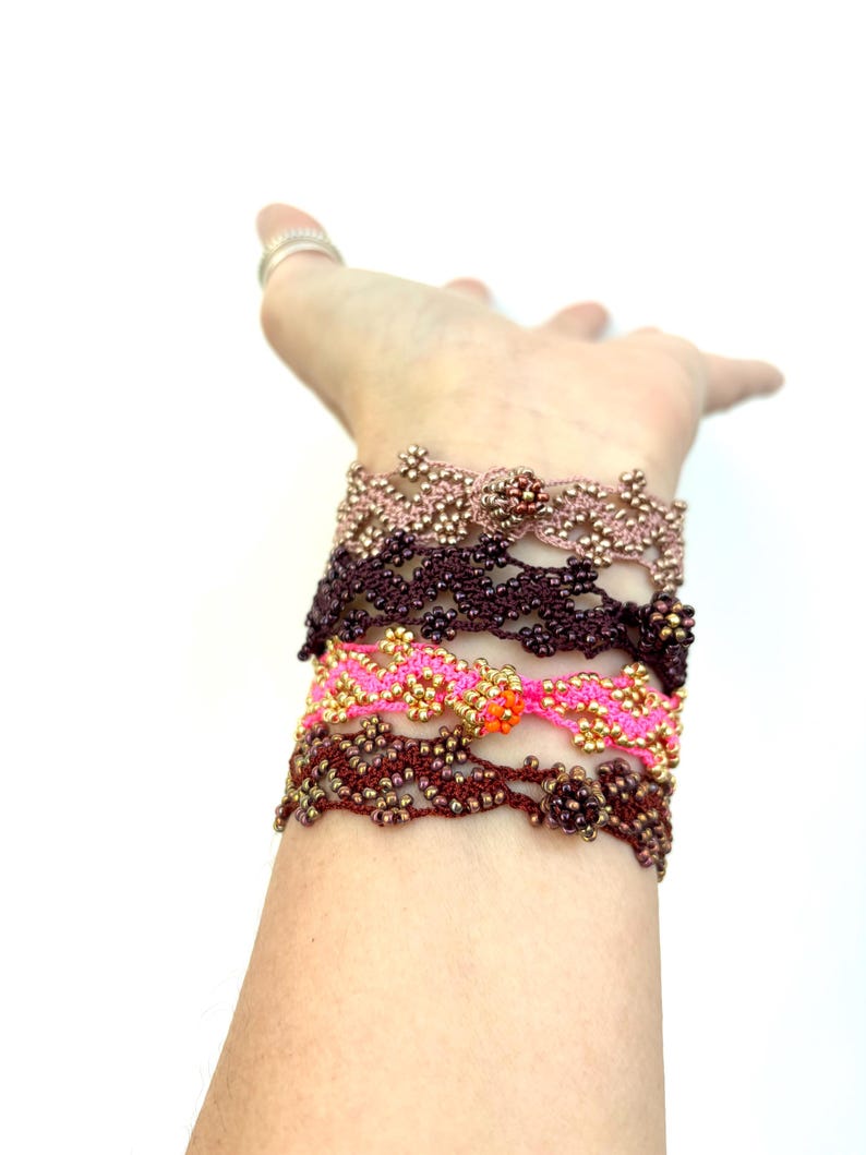 May include: A collection of beaded bracelets in various colors, including pink, brown, and gold. The bracelets are intricately designed with floral patterns and are stacked on a wrist. The background is plain white.
