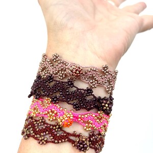 May include: A collection of handmade beaded bracelets in various colors, including pink, brown, and gold. The bracelets feature intricate floral patterns and are worn stacked on a wrist.