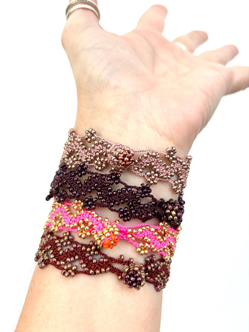 May include: A collection of handmade beaded bracelets in various colors, including pink, brown, and gold. The bracelets feature intricate floral patterns and are worn stacked on a wrist.