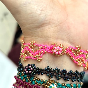 May include: Close-up of several beaded bracelets in various colors. The top bracelet is bright pink with gold beads. Below are bracelets in black, teal, and magenta, all with floral patterns. The bracelets are handmade.