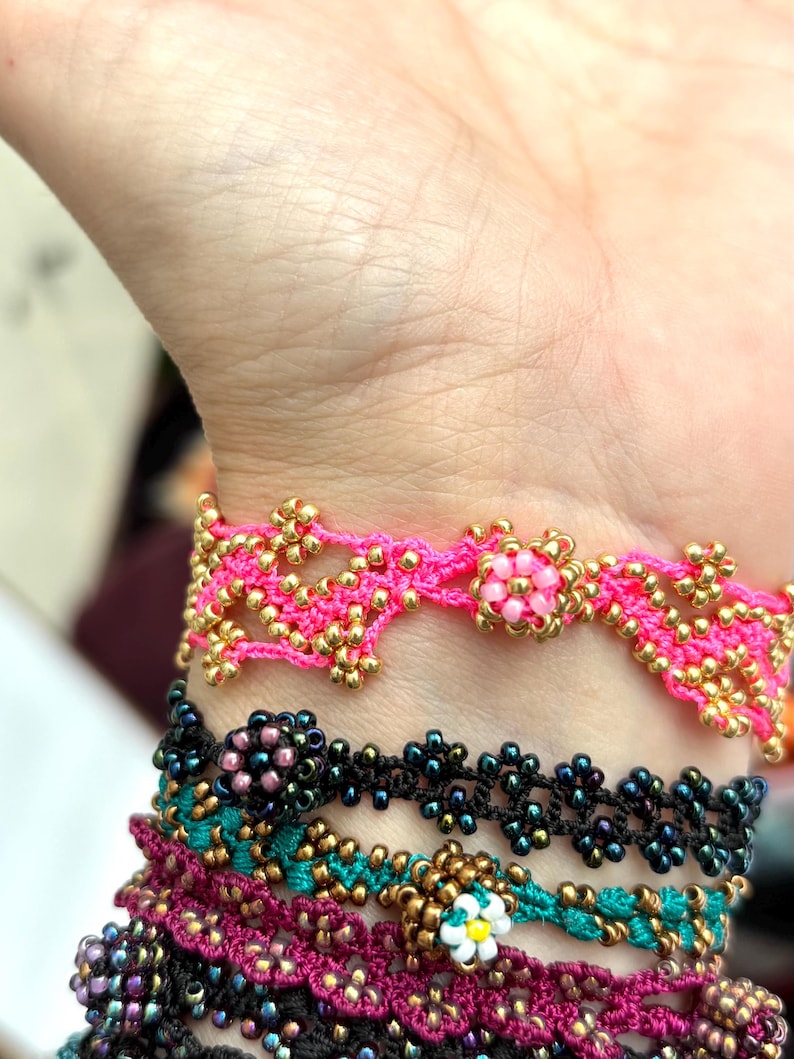 May include: Close-up of several beaded bracelets in various colors. The top bracelet is bright pink with gold beads. Below are bracelets in black, teal, and magenta, all with floral patterns. The bracelets are handmade.