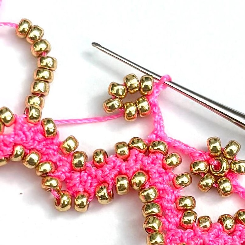 May include: Close-up of pink crochet work with gold beads. A silver crochet hook is inserted into the pink thread, showing the process of adding the beads. The beads are small and round, adding texture and detail to the craft.