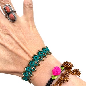 Op de afbeelding: Een hand met een zilveren ring met twee oranjerode stenen. Een turquoise kanten armband met gouden kralen en een zwarte armband met een roze roos en groene bladeren zijn ook zichtbaar. Een derde armband met bruine kralen is ook afgebeeld.