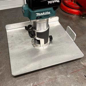 Op de afbeelding: Een teal en zwarte Makita 18V 5.0Ah accu-freesmachine gemonteerd op een zilveren metalen basis. De freesmachine heeft een zwarte accu met de tekst "Makita 18V Lithium-ion 5.0Ah" erop gedrukt.