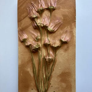 May include: A rectangular wall hanging featuring a relief of stylized flowers. The flowers are pink with gold accents, set against a textured, tan background. A small, gold-colored metal loop is at the top for hanging.