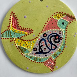 May include: A round, ceramic ornament with a painted bird design. The bird features pink, yellow, blue, and white details, outlined in red dots, against a light green background. A small hole is at the top for hanging.