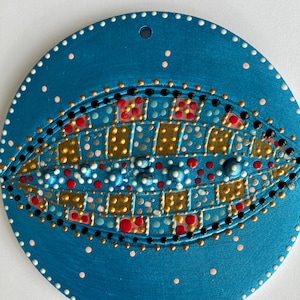 May include: A round, teal-colored decorative piece with an eye-like design. The eye is detailed with gold, red, black, and white dots, creating a textured appearance. The outer edge has white dots, and a hole is at the top.
