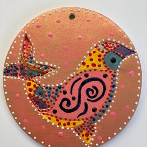 May include: A round, hand-painted wooden ornament featuring a stylized bird design. The bird is primarily pink with yellow, blue, and red accents, and black swirl details. The background is a salmon color with small pink dots and white dots around the edge.