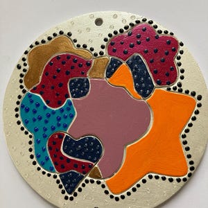 May include: A round, white, painted ornament with a hole at the top for hanging. The ornament features a colorful abstract design with shapes in shades of orange, pink, red, blue, gold, and black.