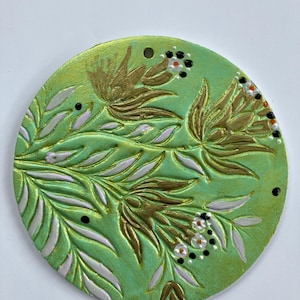 May include: A round, green decorative piece with a floral design. The design features gold-colored flowers and leaves, with white and black accents. The piece has a textured surface and a hole at the top.