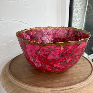 Könnte beinhalten: Eine dekorative Schale mit leuchtend rosa Innenraum und goldfarbenem Rand. Die Schale weist ein Blumenmuster in Grüntönen auf und steht auf einem hellbraunen Holzständer. Das Design der Schale verleiht einen Hauch von Eleganz.