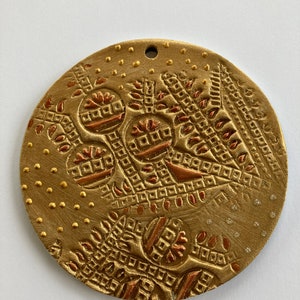 May include: A round, gold-colored ornament with a textured surface and a copper-colored design. The ornament has a hole at the top for hanging.