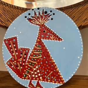 May include: A round, hand-painted decorative plate featuring a stylized red bird design on a light blue background. The bird has a triangular body and arrow-like tail, with gold, white, and green accents. The plate is trimmed with white dots.