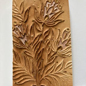 Könnte beinhalten: Eine Terrakotta-Fliese mit einem erhabenen Blumendesign in Gold und Weiß. Die Fliese hat einen rustikalen, handgefertigten Look und wäre eine schöne Ergänzung für jedes Zuhause.
