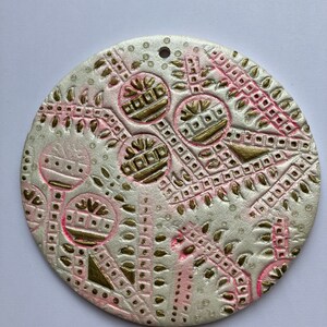 May include: A round, white ceramic ornament with a pink and gold design. The ornament features a repeating pattern of leaves, circles, and geometric shapes.