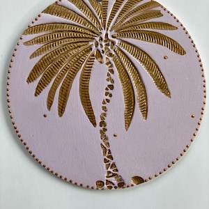 Puede incluir: Un adorno de pared redondo de cerámica de color lavanda con un diseño de palmera dorada. La palmera está en el centro, con un tronco texturizado y hojas detalladas. El borde está salpicado de pequeños detalles dorados.