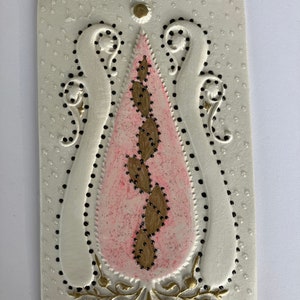 Könnte beinhalten: Eine weiße Keramikplatte mit einem erhabenen Design in Form eines rosa und goldenen Tränen tropfens mit schwarzen Punkten. Der Tränen tropfen ist von einem weißen, erhabenen, verzierten Design mit schwarzen Punkten und goldenen Akzenten umgeben.